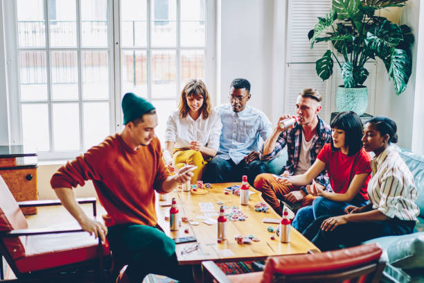 Diverse cheerful hipster guys drinking beverage and playing cards at table with chips for poker in modern apartment.Positive casual dressed multicultural friends spending free time together in flat Diverse cheerful hipster guys drinking beverage and playing cards at table with chips for poker in modern apartment.Positive casual dressed multicultural friends spending free time together in flat board game photos stock pictures, royalty-free photos & images