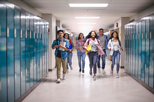 Cheerful male and female students running in illuminated corridor. Full length of happy teenage friends are amidst lockers. They are at high school.