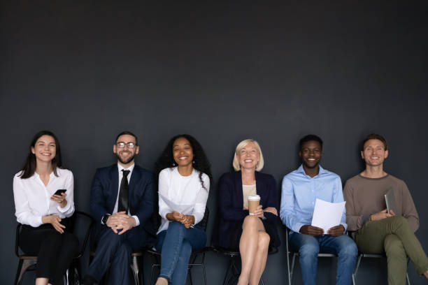 Diverse successful staff seated on chairs smiling looking at camera Near black wall studio background sitting six diverse happy entrepreneurs, successful corporate staff members, businesspeople ready for negotiation, wait for job interview sit in row queue, hr concept candidate stock pictures, royalty-free photos & images