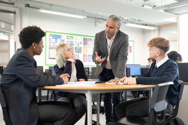 Diverse teenage students listening to mature male educator Male and female classmates in school uniforms sitting at round table in collaborative workspace and interacting with teacher supervising their discussion. british curriculum schools stock pictures, royalty-free photos & images