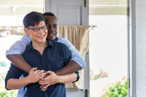 Diversity gender of LGBTQ gay couple between Asian and African ethnicity hugging each other at home for same sex marriage and pride month concept stock photo