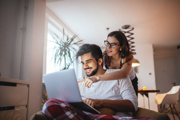 Do the little things that make each of you smile Shot of a happy young couple embracing while using laptop at home. laptop couple stock pictures, royalty-free photos & images