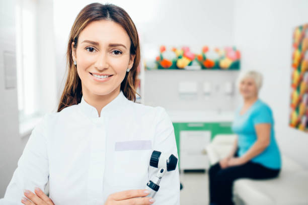 Doctor dermatologist with dermatoscope, smiling while looking at camera. Behind her sitting senior patient. Doctor dermatologist with dermatoscope, smiling while looking at camera. Behind her sitting senior patient. dermatologist stock pictures, royalty-free photos & images
