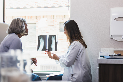 An orthopedic doctor examines a senior woman's bone fracture during a medical appointment. The doctor is reviewing an x-ray image of the woman's foot with the patient.