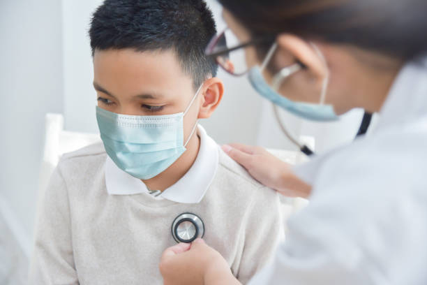 Doctor examining Asian boy wearing protective face mask with stethoscope stock photo