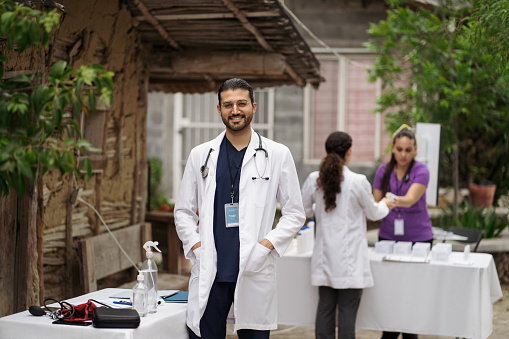 A young latin male doctor standing next to the consultation table in a rural area and smiling at the camera.