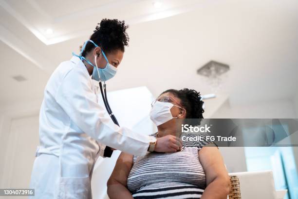 Doctor listening to patient's heartbeat during home visit - wearing face mask