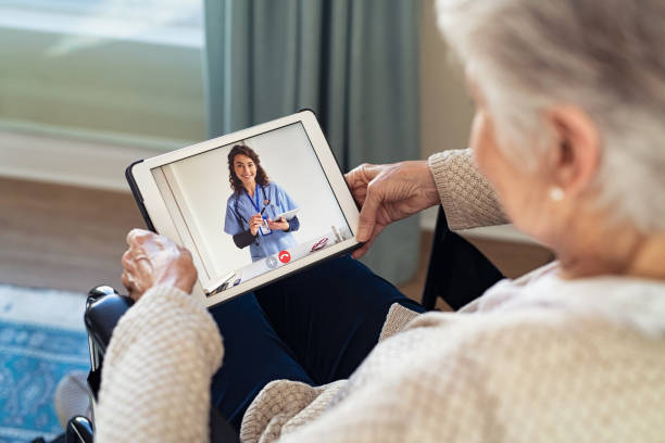 Doctor on video call with disabled senior patient Back view of senior woman sitting in wheelchair making video call with her doctor while staying at home during covid pandemic. Close up of helpful general practitioner in video conference with old woman on digital tablet. Sick woman in online consultation from home: distance and telehealth concept. nurse talking to camera stock pictures, royalty-free photos & images