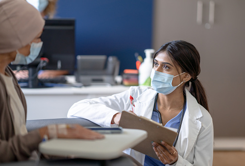 A female doctor bend's down to eye level as she talks with a cancer patient who is receiving her treatment intravenously.  She is wearing a white lab coat and has a clipboard in hand as she takes notes.  The patient is dressed casually and she sits back in the chair waiting for her treatment to finish, and both are wearing medical masks to protect them from COVID.