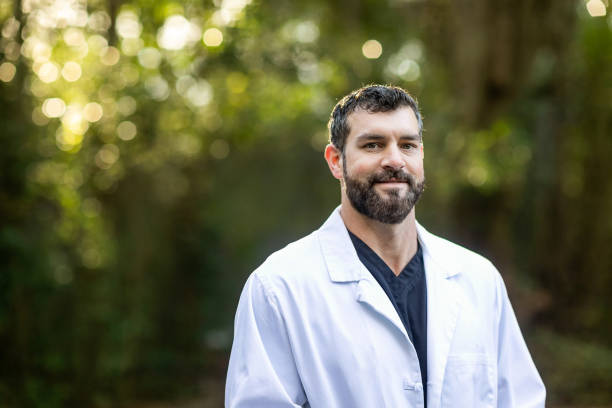A doctor with dark hair and a beard in a white lab coat standing outside in a natural green environment stock photo