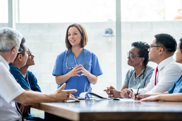 Doctors Meeting A multi-ethnic group of medical staff are indoors in a hospital. They are wearing medical clothing. A Caucasian female doctor is giving a presentation to the others. medical occupation stock pictures, royalty-free photos & images