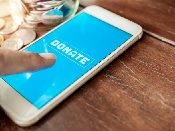 Donation Online Finger pressing donate icon button on blue screen on white mobile phone with coins money on wooden background with copy space. Donation online concept. charitable donation stock pictures, royalty-free photos & images