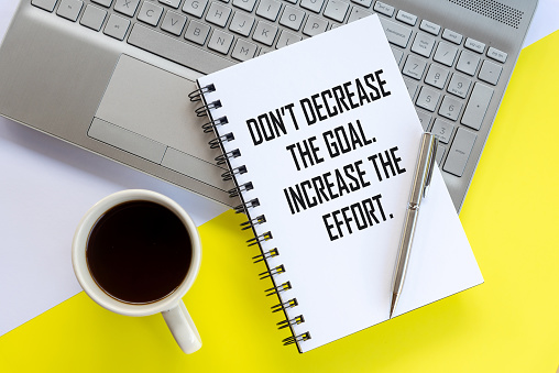 Business motivational words on a book - Don't decrease the goal. Increase the effort. A flat lay concept with cup of coffee, a book, pen and laptop on white and yellow table background.