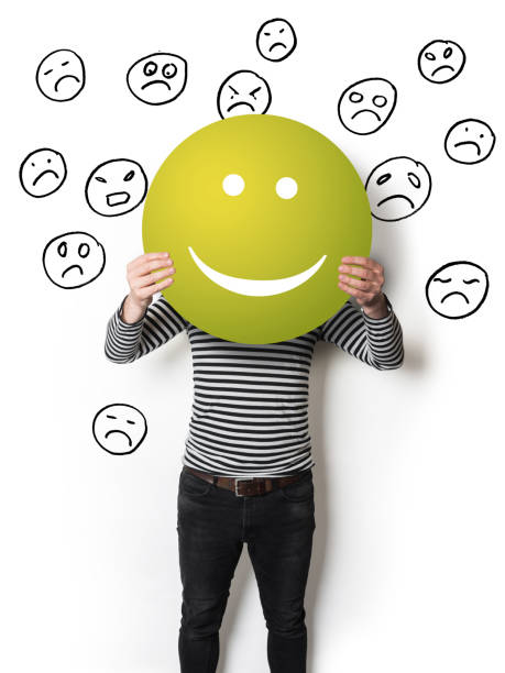 Man holding a green smiling emoticon in front of sad faces hand drawn on a white wall.