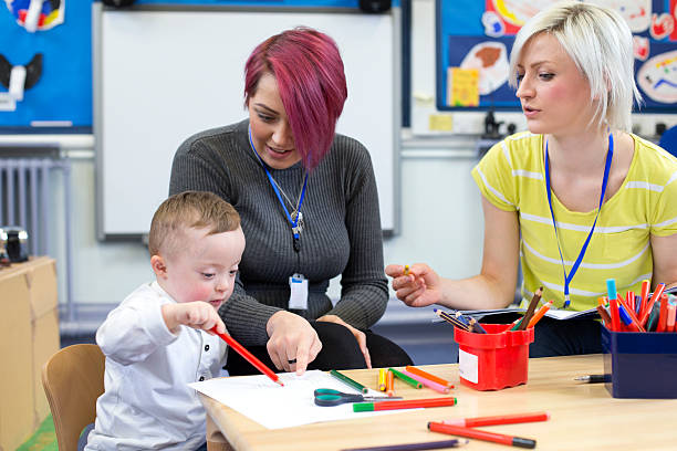 Down Syndrome boy at Nursery Nursery teacher sitting with a parent and her Down Syndrome son in the classroom. they are discussing the little boys progress. preschool teacher stock pictures, royalty-free photos & images
