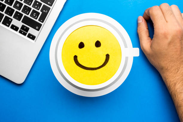 tekening van een gelukkige glimlachende emoticon op een geel document en blauwe achtergrond met laptopcomputer en hand die over bureau rust. - happy friday emoticon stockfoto's en -beelden