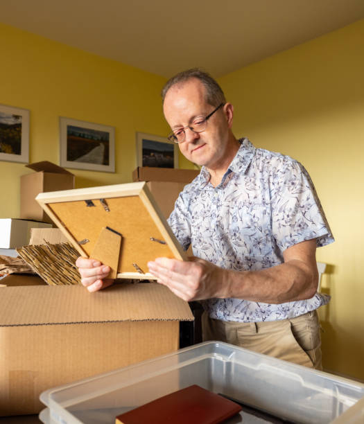 Dreaming about an old picture, a senior man alone in empty apartment moving house, packing stuff in cardboard boxes looking at nostalgic items stock photo