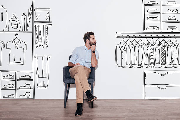 Dreaming about new wardrobe. Young handsome man keeping hand on chin and looking away while sitting in the chair against illustration of closet in the background day dreaming photos stock pictures, royalty-free photos & images