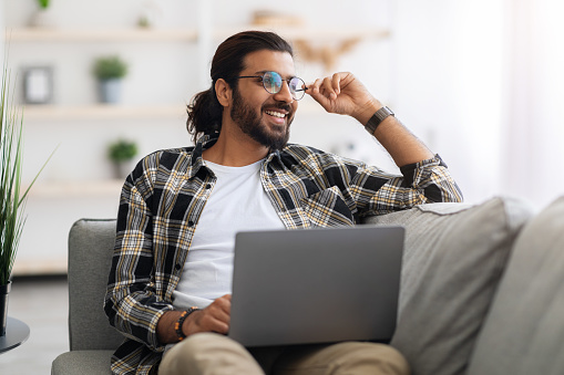 Dreamy arab guy freelancer working from home, wearing glasses, sitting on couch, using laptop and looking at copy space, thinking about career opportunities or looking for creative solution