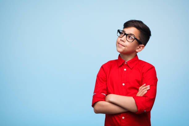 Dreamy boy with arms crossed Adorable young man standing with arms crossed and daydreaming on the blue background. boys glasses stock pictures, royalty-free photos & images