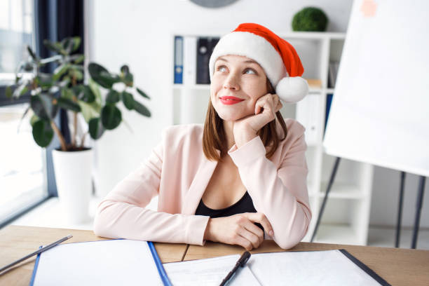 Dreamy positive and happy young woman look left up and smile. Wear red cap or hat. Festive mood. Celebrating new year or Christmas. Alone at work. Sit in office. stock photo