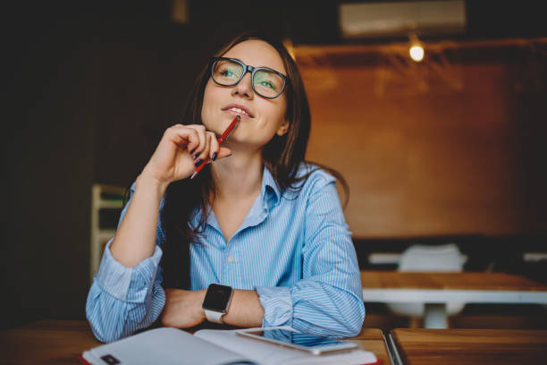 Dreamy woman podring while working on journalistic publication sitting with notebook in cafe,thoughtful female student in eyewear doing homework task solving problems and analyzing information stock photo