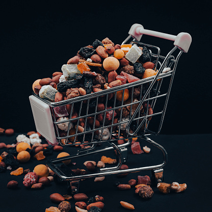 An overflowing shopping cart with a nut mixture scattered on a black background. The concept of discounts and sales on dried fruits.