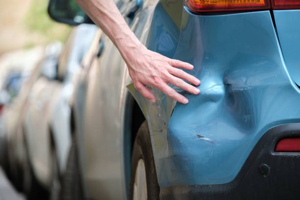 Driver hand examining dented car with damaged fender parked on city street side. Road safety and vehicle insurance concept stock photo