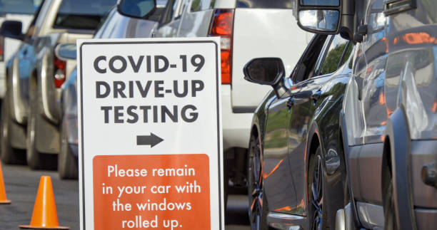 un letrero de "covid-19 drive-up testing" se encuentra en primer plano mientras los coches y otros vehículos esperan en una línea de prueba covid-19 (coronavirus) fuera de una clínica médica/hospital al aire libre (segunda ola) en segundo plano - covid test fotografías e imágenes de stock