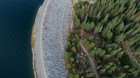 looking straight down on green pine trees and lake divided by a road and rocks. Shot from high in the sky with drone