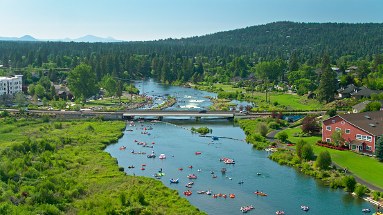 Aerial establishing shot of Bend, Oregon, on a hot and sunny day in summer. Scores of people are tubing, paddleboarding, kayaking and surfung in the water of the Deschutes River, especially around the Whitewater Park, a set of artificial rapids and waves.