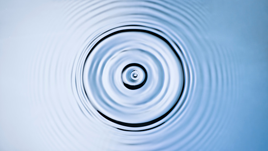 Overhead view of droplet creating a splash and ripples when hitting the blue and silver coloured water surface.