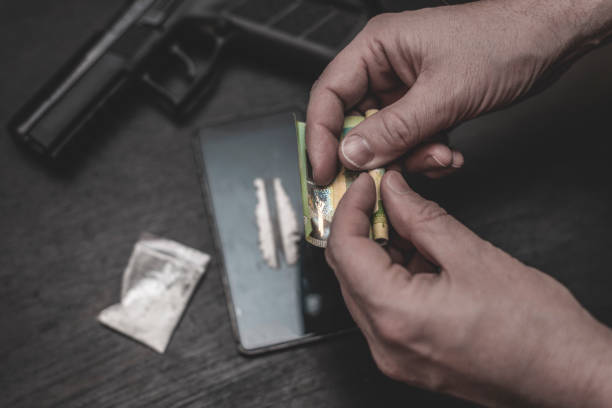 Drug addiction The man rolls the banknote, track of cocaine on a mobile phone, a pistol and a bag of cocaine on the side on the table. mephedrone stock pictures, royalty-free photos & images
