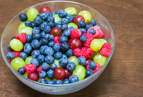 A chilled bowl filled with an assortment of fresh summer fruits including red and green grapes, blueberries and raspberries on a wooden table,