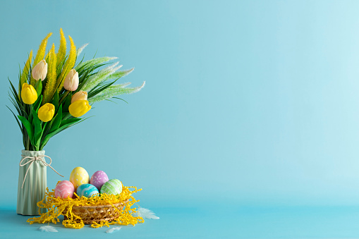 Multi colored hand-painted Easter eggs in a basket with the bouquet of tulips in a vase. Blue background. Space for copy.