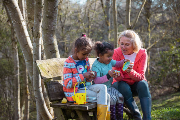 Easter Sunday with Grandma! A grandmother and her two young granddaughters sitting on a wooden bench in a woodland area in Hexham, Northumberland. They have collected easter eggs on an Easter egg hunt, they have their baskets and they're looking at all the eggs they've found. easter sunday stock pictures, royalty-free photos & images