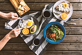 Top view shot of breakfast table. Poached egg with bread, sliced orange and avocado are on plates. Coffee mugs, cheese plate and salad with avocado, tomatoes and arugula are on the table with a grey tea towel. Hand slicing egg on the plate.