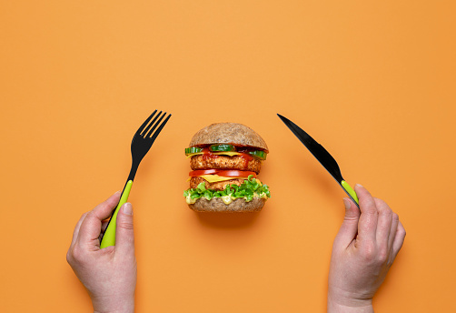 Woman eating vegan burger top view on a colored table. Woman hands holding knife and fork, preparing for eating a vegan sandwich.