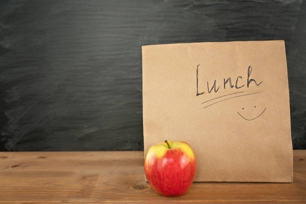 Eco friendly brown paper lunch bag on wooden table with red apple. Chalk board on background. Back to school concept stock photo