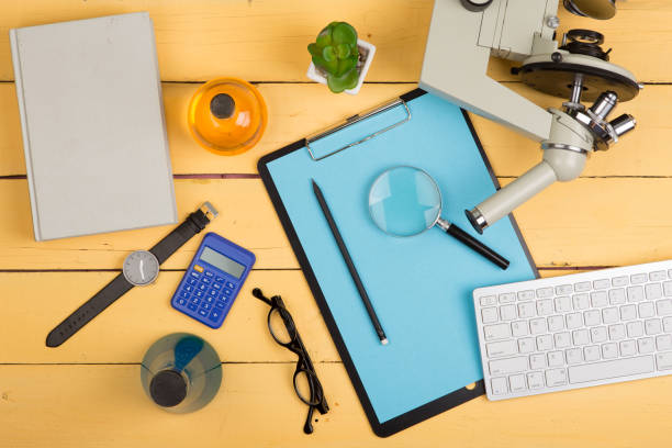 Education and science concept - microscope, book, magnifying glass, calculator, watch, blank clipboard, computer keyboard, eyeglasses and chemical liquids stock photo