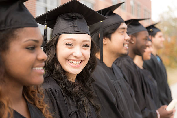 Education: Graduates stand in row on college campus. Six multi-ethnic friend graduates excitedly wait for their name to be called during graduation ceremony. Mixed-race girl looks back at camera. School building background. best schools stock pictures, royalty-free photos & images