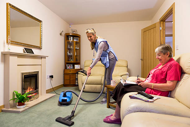 Elderly Care in the Home Care worker making a home visit. Female carer is hoovering the living room to help an elderly woman. The woman is sitting on a sofa relaxing hovering stock pictures, royalty-free photos & images
