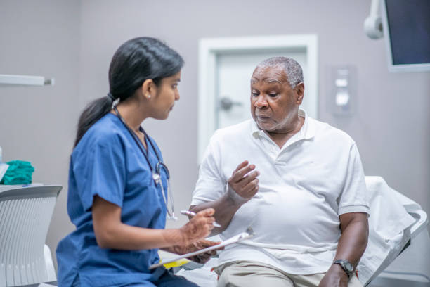 Elderly Male Patient Talking with Female Doctor stock photo Elderly African American male patient sits across from a female doctor during Covid-19 outbreak.  The doctor is taking notes on his patient chart. medical research stock pictures, royalty-free photos & images
