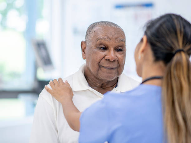 Elderly Man Visits Doctor stock photo An elderly African American man sits across from a female doctor during a visit.  The Female doctor is wearing blue scrubs and has her back to the camera. sad old black man stock pictures, royalty-free photos & images