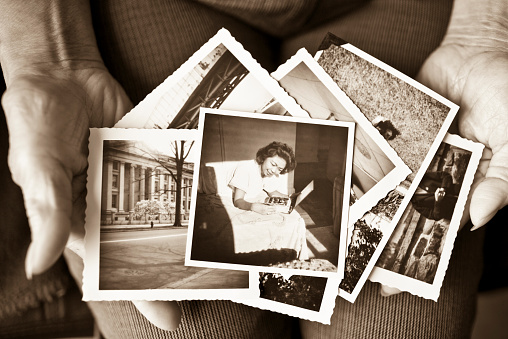 Toned image of an elderly, senior woman holding old vintage photographs of herself and of other places in her hands, showing her sentimental memories, past, and places travelled.  Only her hands are shown in the image