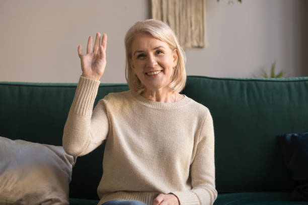 Elderly woman makes video call waving hand greets friend Attractive elderly woman sitting on couch at home looking at camera makes video call waving hand greeting friends or relatives, communication distantly, modern wireless technologies easy usage concept adult cam rooms stock pictures, royalty-free photos & images