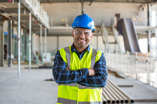 Electrician wearing work helmet, protective eyeglasses and reflective vest standing at the construction site with arms crossed, smiling at camera. Man at work.