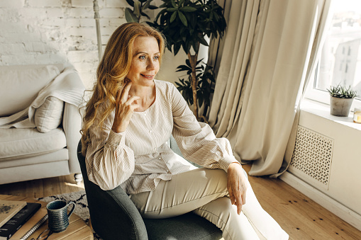 Elegant, charming, beautiful mature woman sitting in lovely stylish chair posing in surroundings of modern living-room, resting after reading favorite book, dressed in striped blouse and suit pants