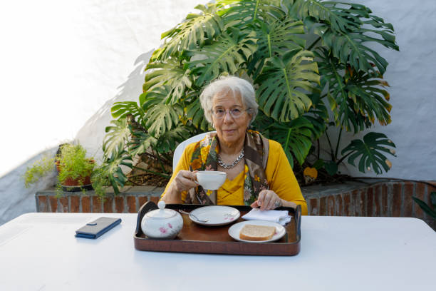Elegant elderly lady, drinking tea in the courtyard. In the background flower bed with large green leafy plant, Monstera deliciosa. A Elegant elderly lady, drinking tea in the courtyard. In the background flower bed with large green leafy plant, Monstera deliciosa. south american woman stock pictures, royalty-free photos & images