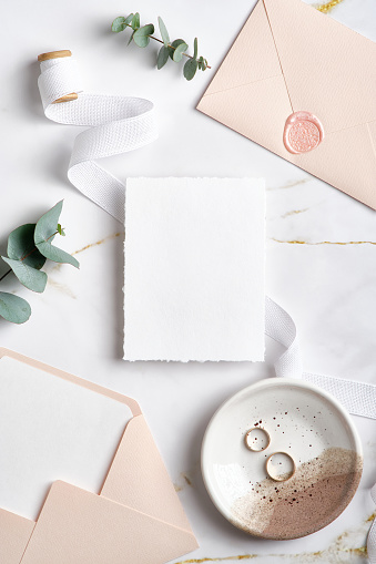 Elegant wedding stationery top view. Flat lay blank paper card mockup, pink envelopes, eucalyptus branches, golden rings on marble background.